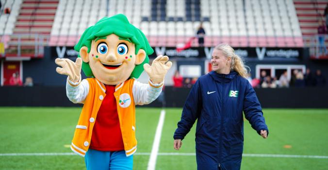 Mascotte Mattie wandelt op een grasveld samen met een vrouw