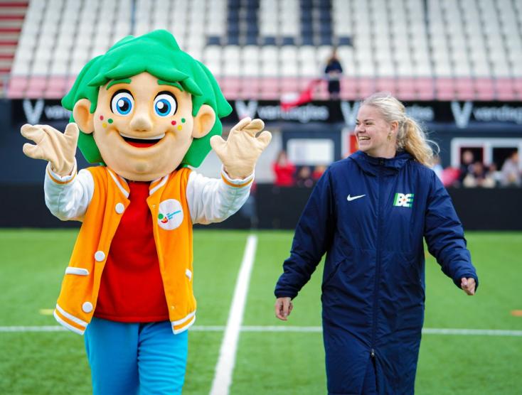 Mascotte Mattie wandelt op een grasveld samen met een vrouw